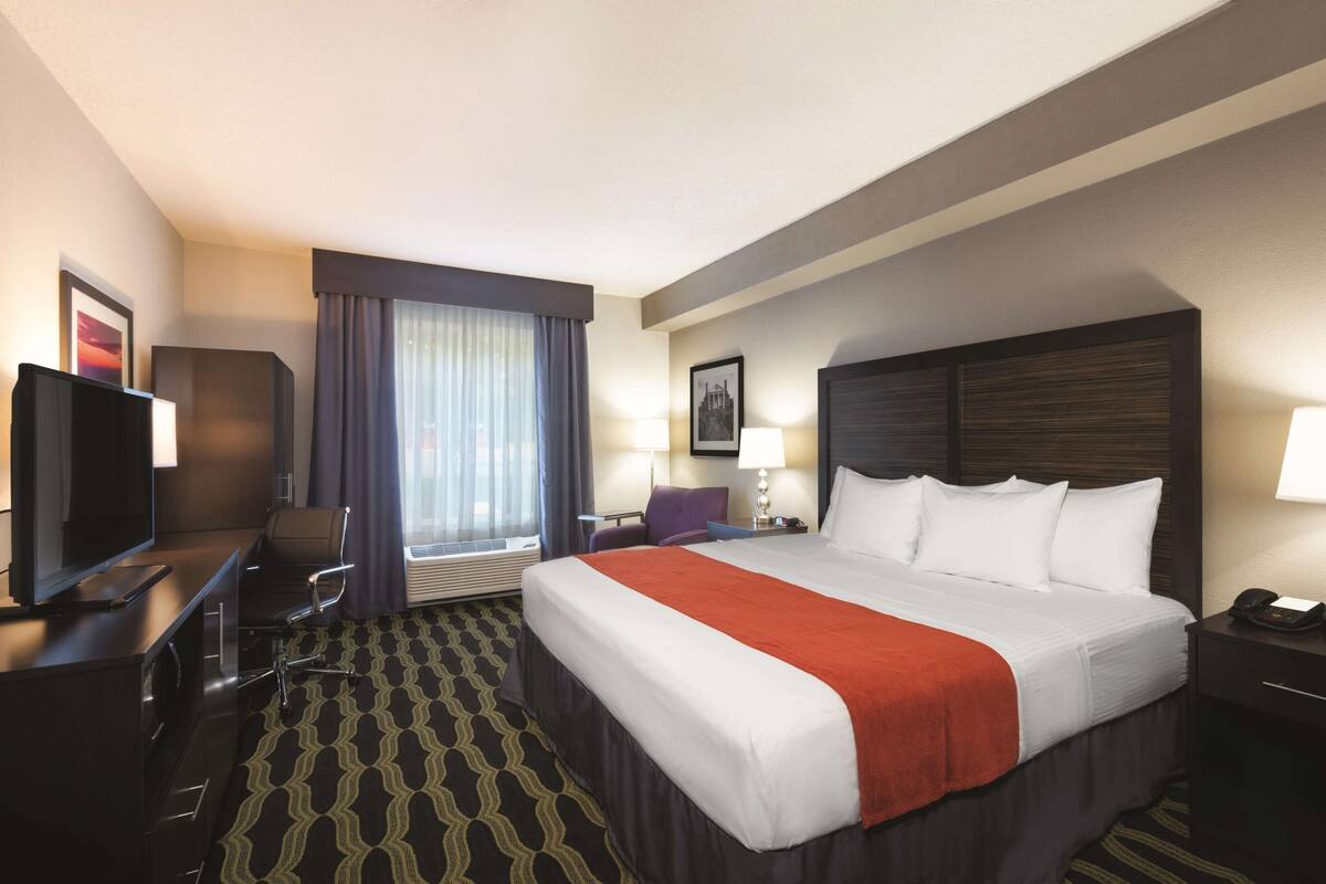 A hotel room with a large bed, a desk with a TV, a chair, a desk lamp, and a window with curtains.