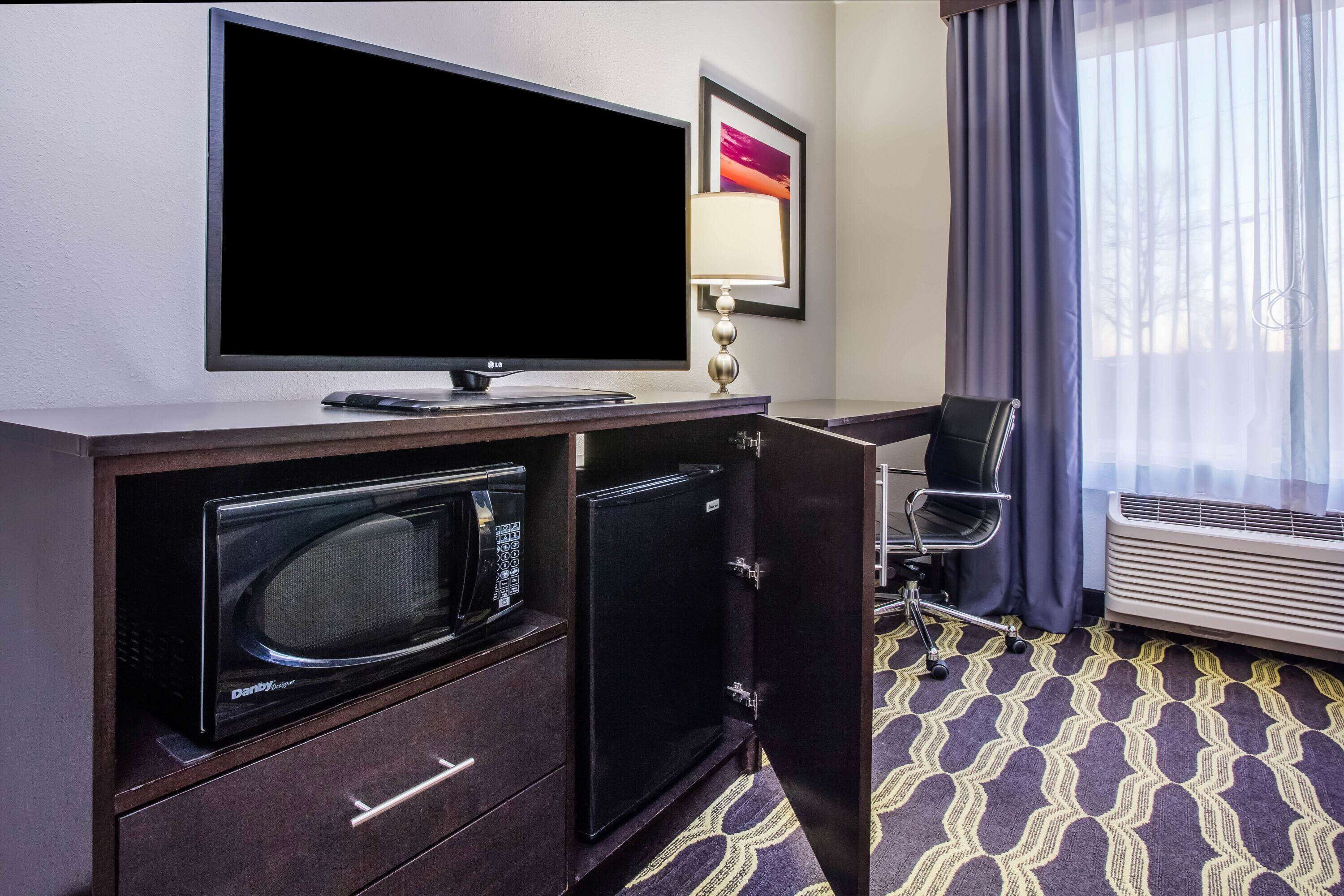 Habitación de hotel con televisor de pantalla plana, microondas, mini refrigerador, escritorio con lámpara, silla y un ventanal con cortinas.