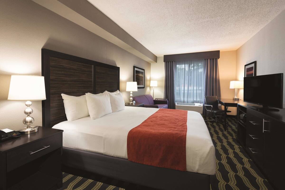 A hotel room with a large bed, two bedside tables with lamps, a desk with a chair, a television, and a window with curtains.