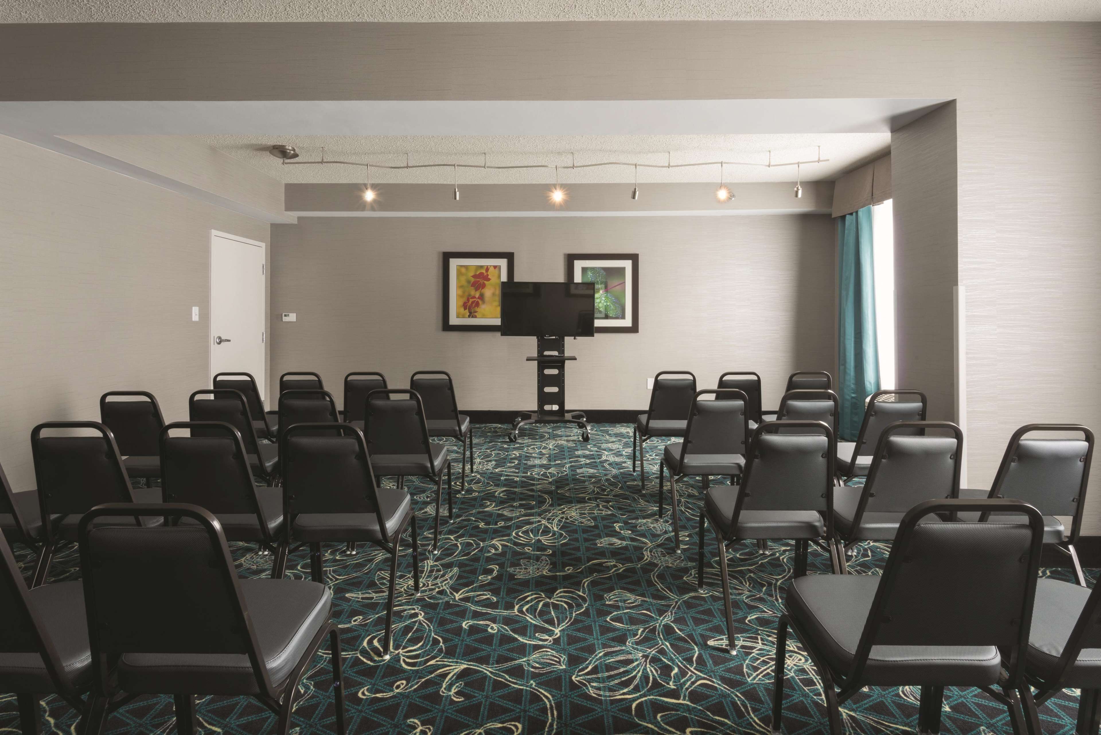 Un salón de conferencias con filas de sillas negras, una pantalla grande y alfombra estampada.