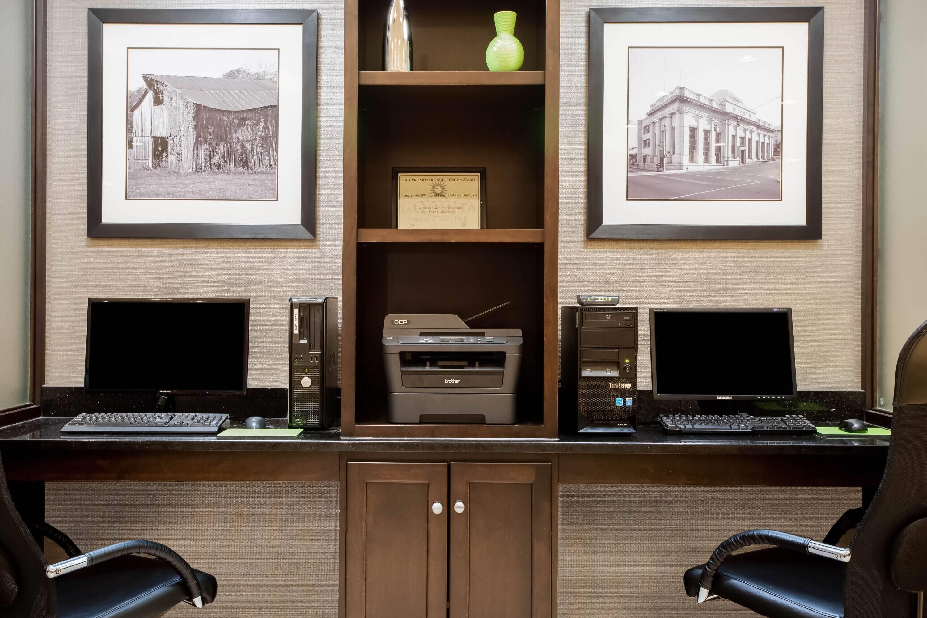 A home office setup with two desks, each with a computer, keyboard, and monitor. There are framed pictures on the wall, a printer, and a cabinet with a door.