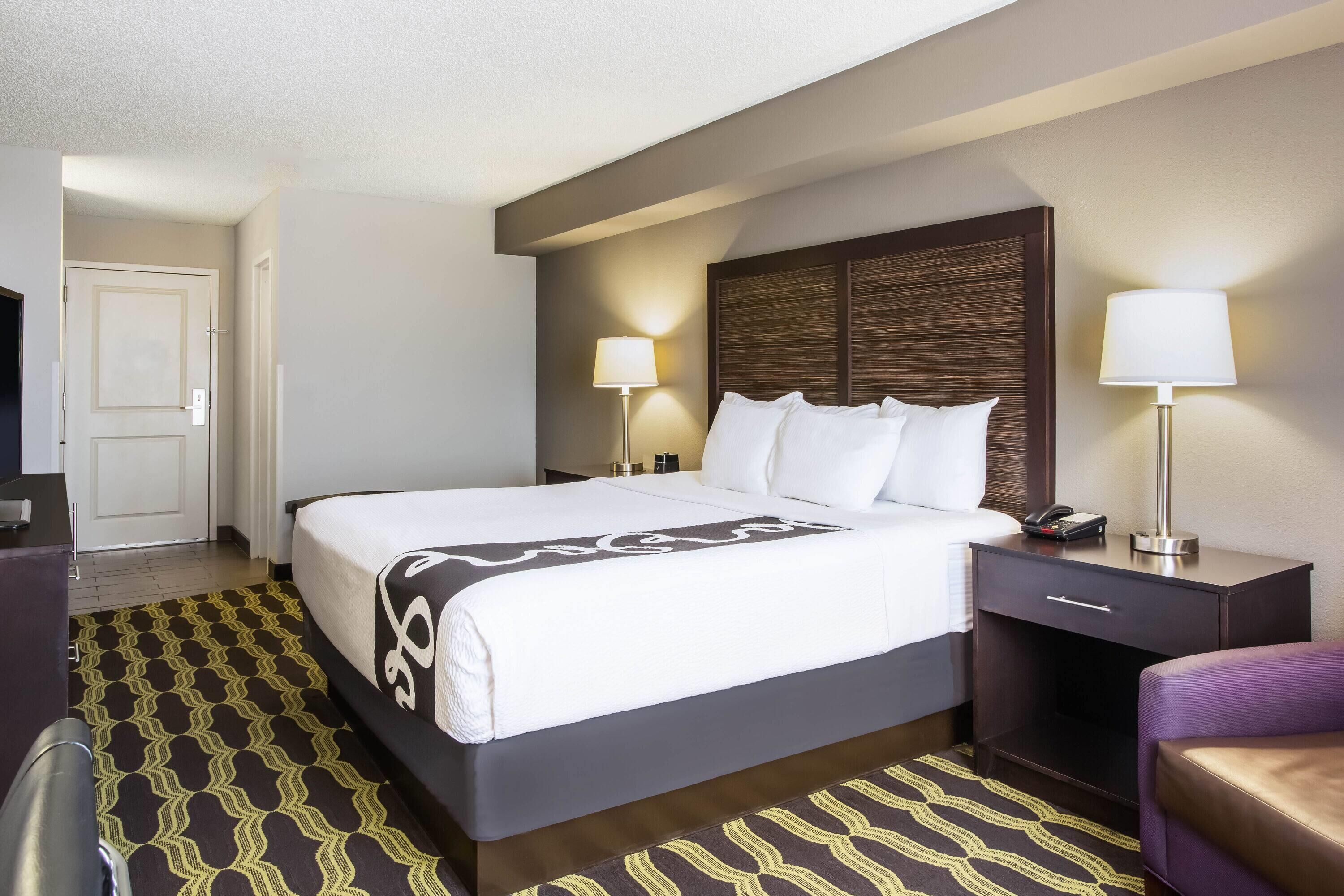 A hotel room with a bed, bedside lamps, a desk with a phone, and a patterned carpet.