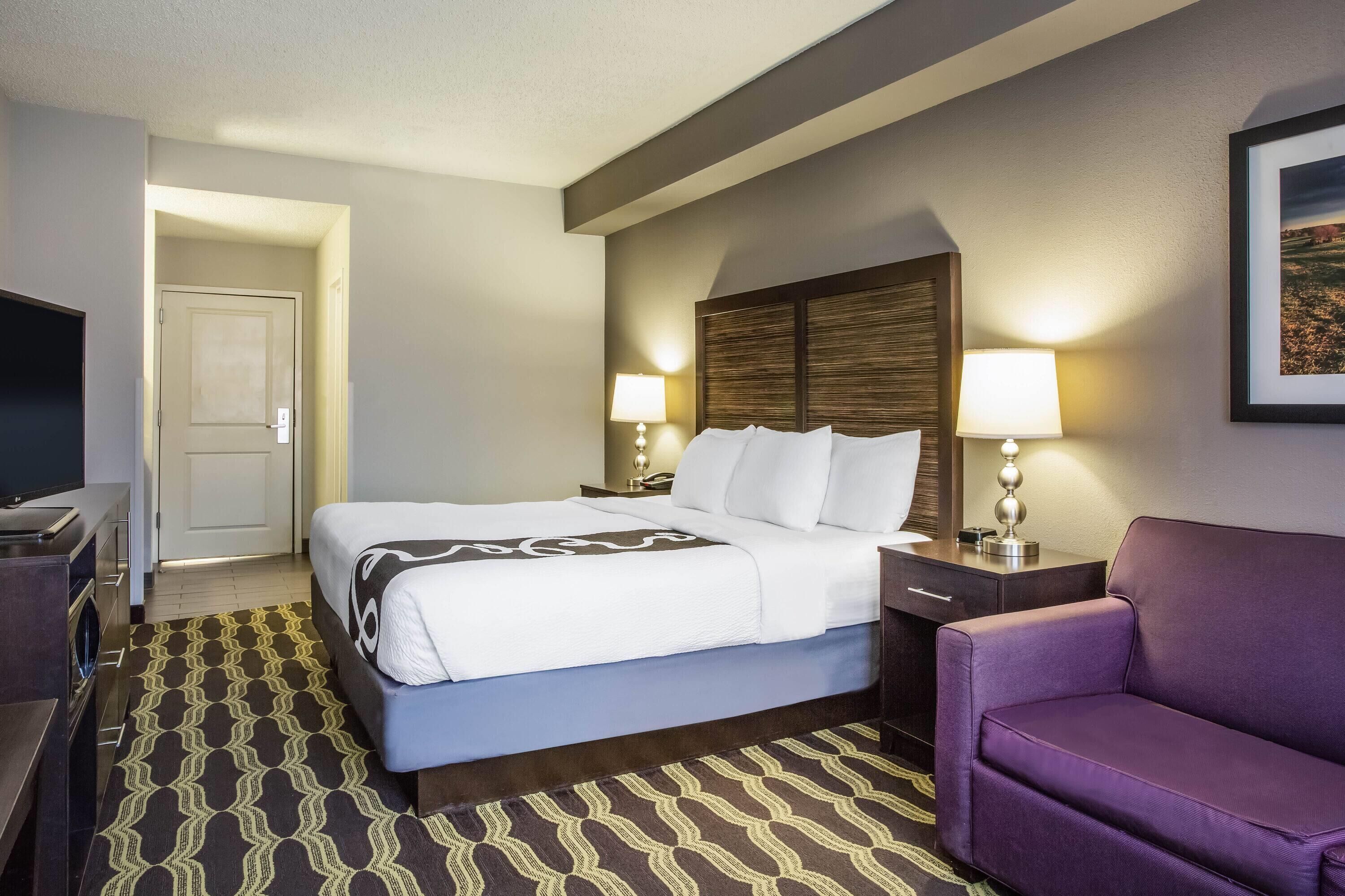 A hotel room with a bed, bedside tables, a television, a purple armchair, and a patterned rug.
