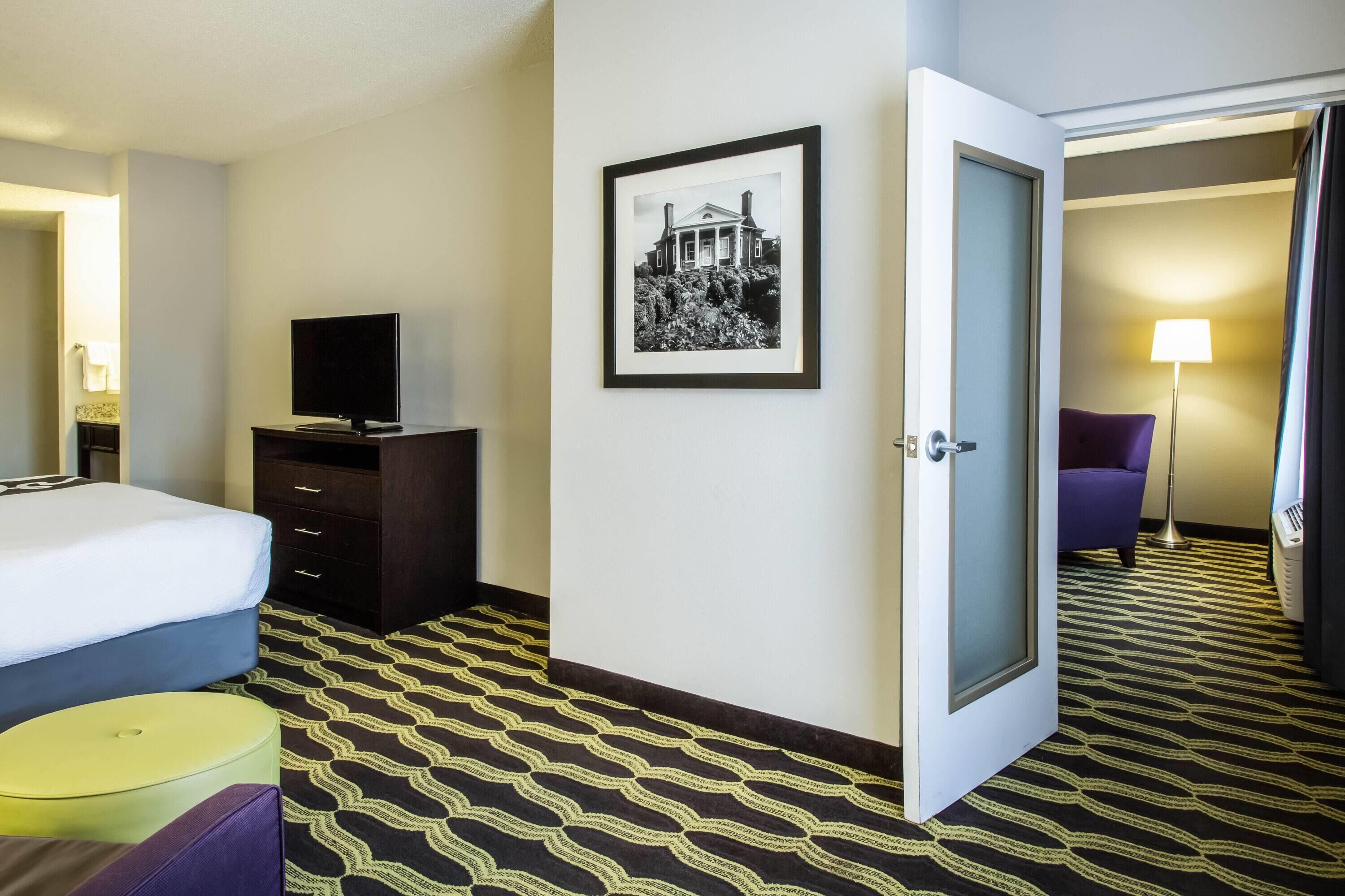 Una habitación de hotel con una cama, un aparador, un televisor, un cuadro enmarcado en la pared y una puerta que da a otra habitación.