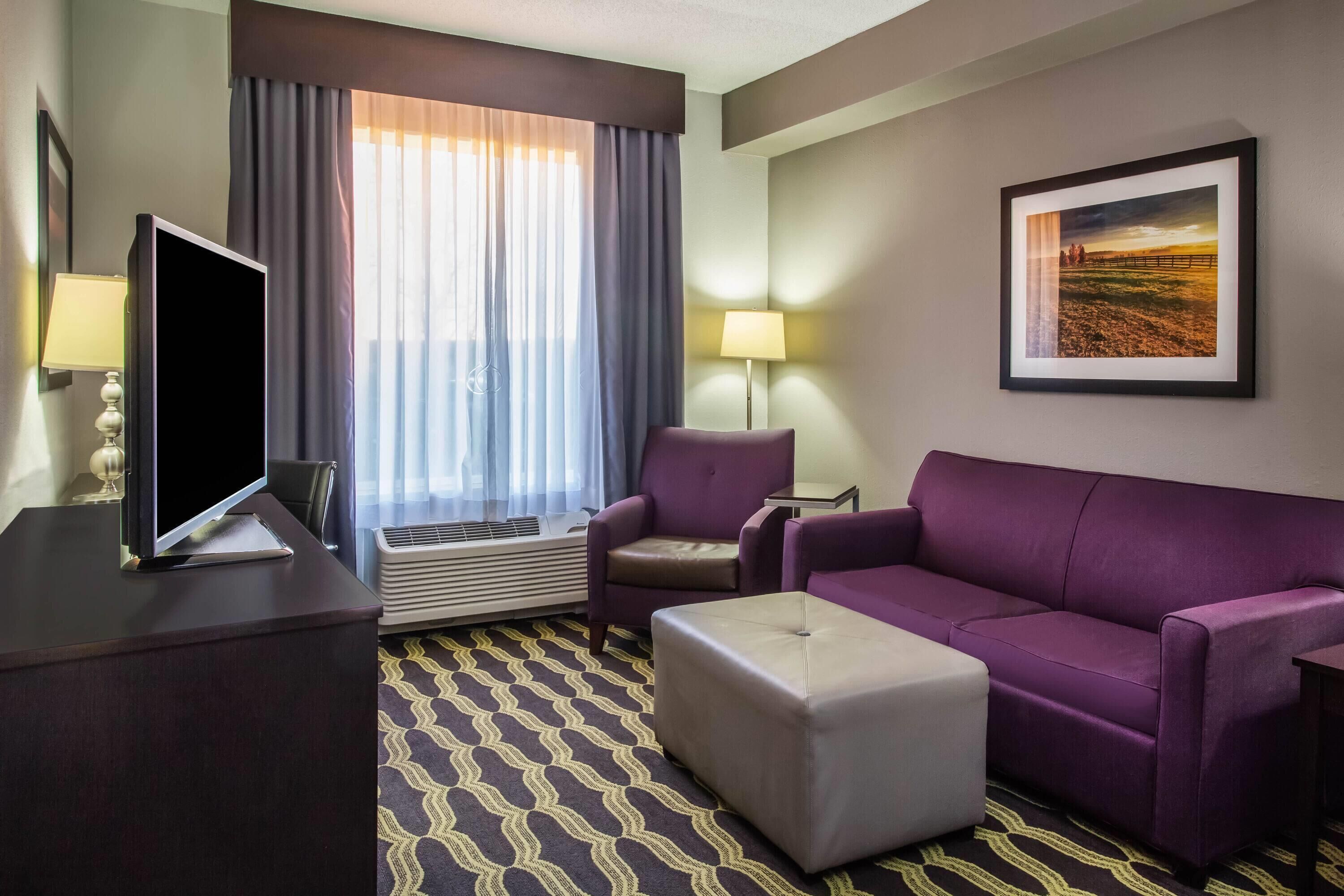 Une chambre d’hôtel avec un bureau, un canapé violet, un pouf, une lampe sur pied et une grande fenêtre avec des rideaux.