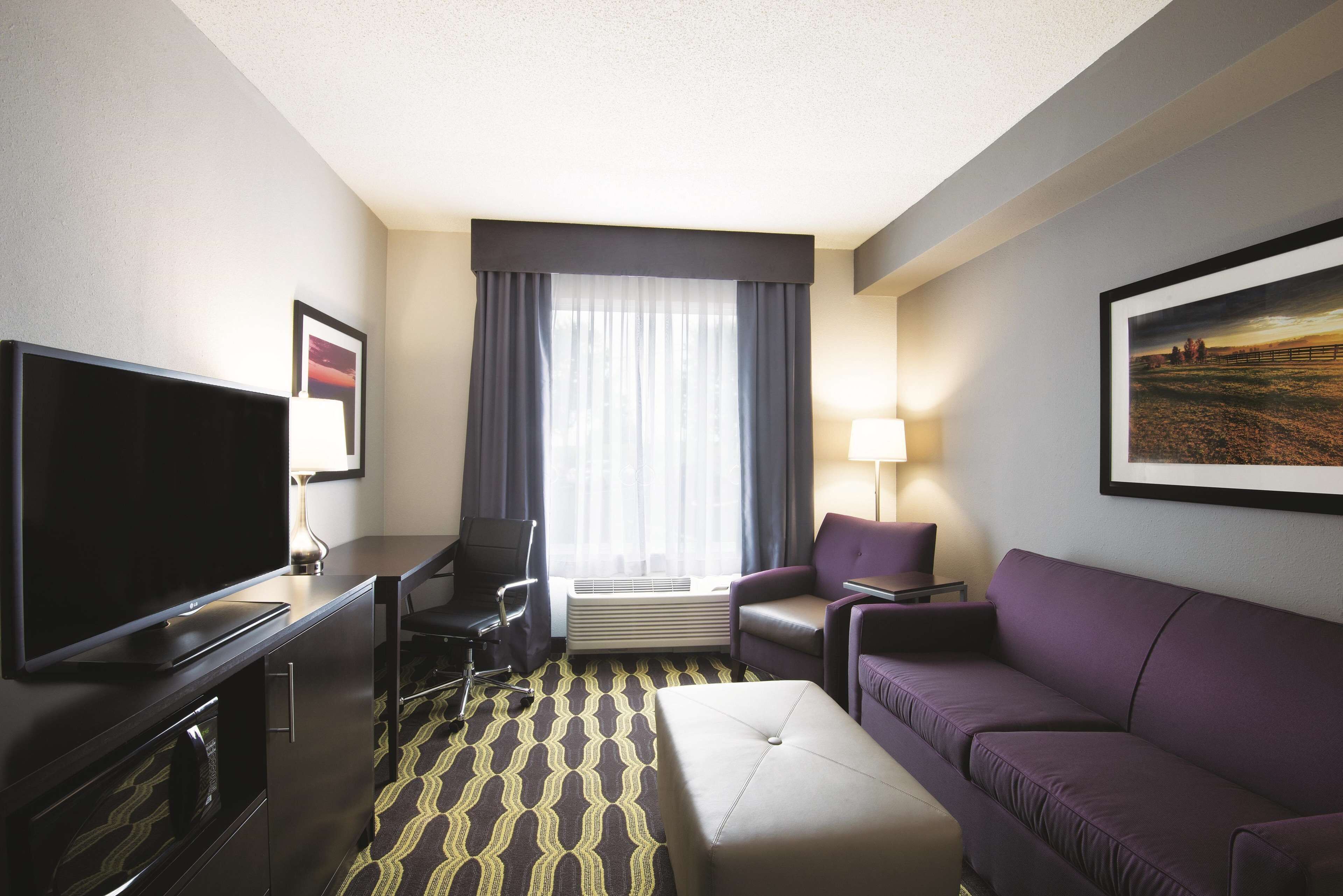 A hotel room with a flat-screen TV, a desk, a chair, a sofa, an ottoman, a lamp, and a large window with curtains.