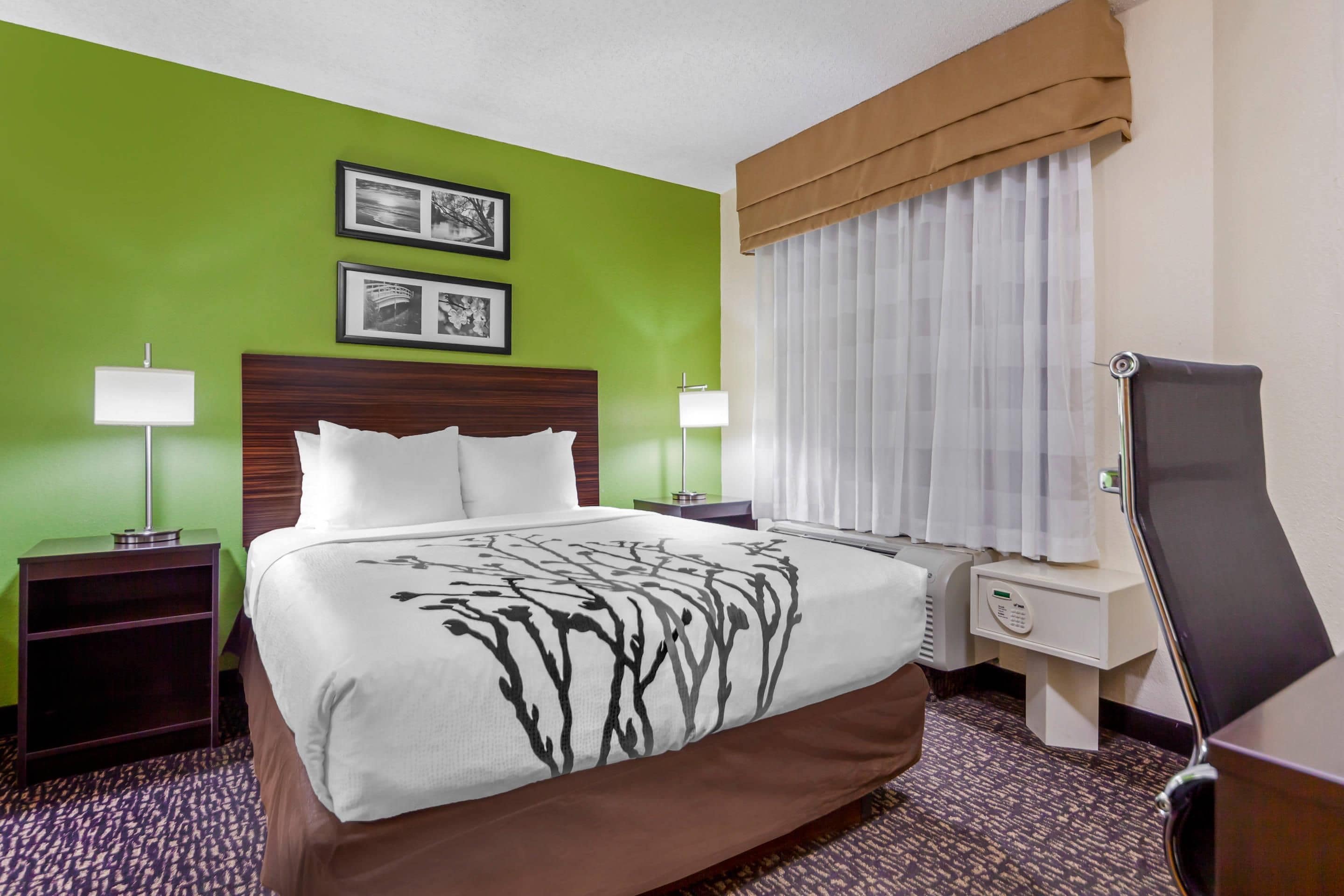A hotel room with a large bed, two bedside tables with lamps, a headboard, a window with curtains, and a wall-mounted TV.