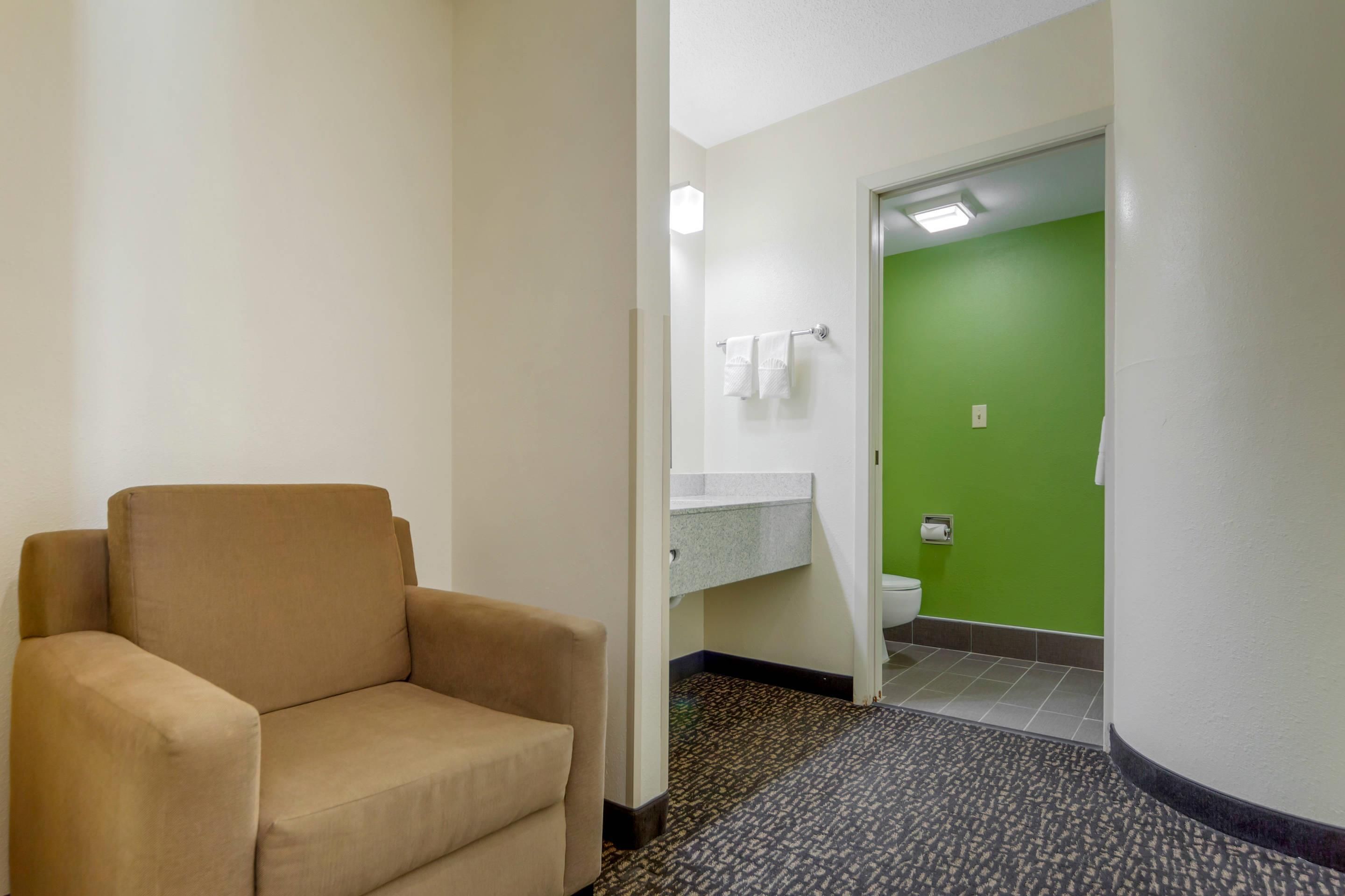 Une chambre d’hôtel avec un fauteuil beige, un petit lavabo avec un miroir et un mur vert.