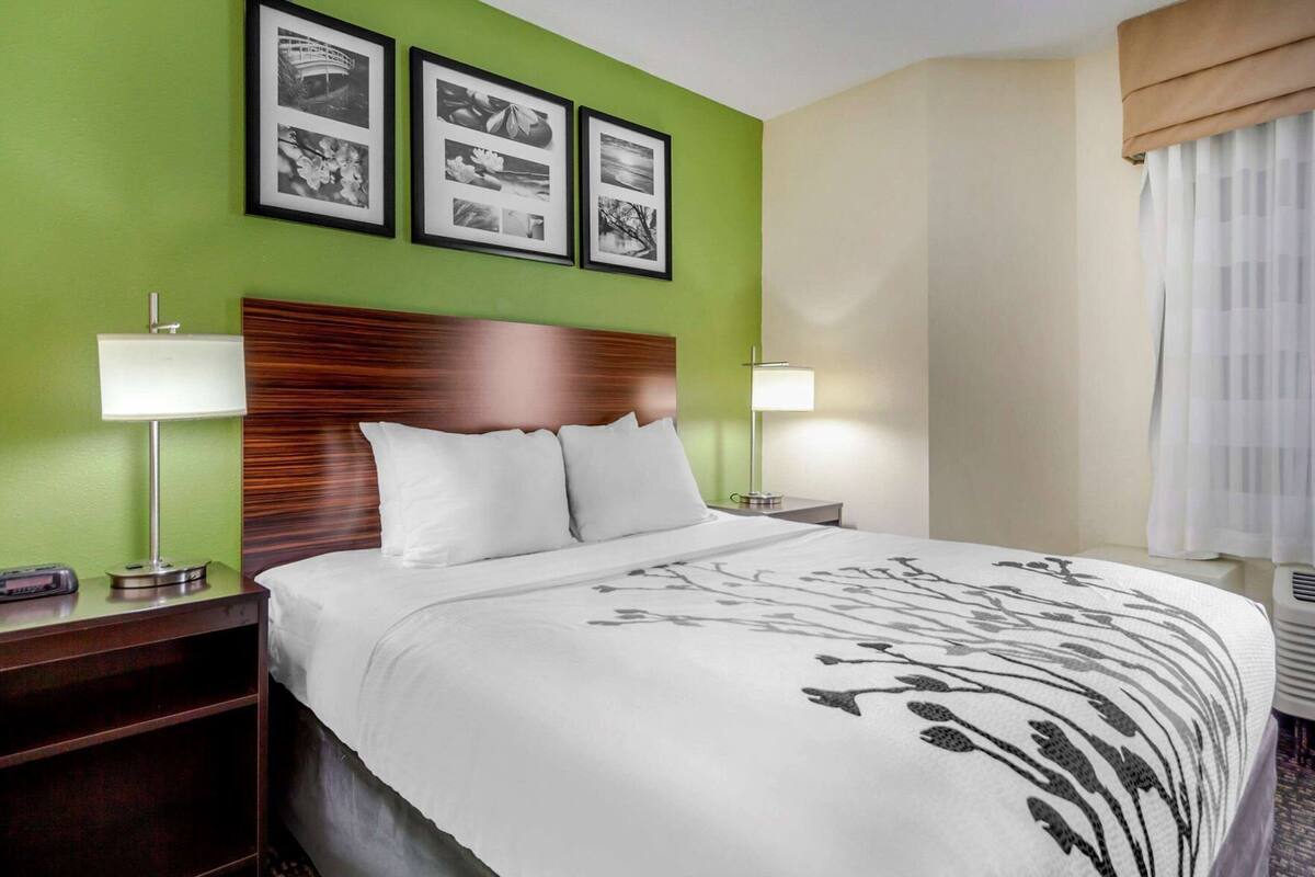 A hotel room with a bed, two bedside lamps, a nightstand, a wall with framed pictures, and a window with curtains.