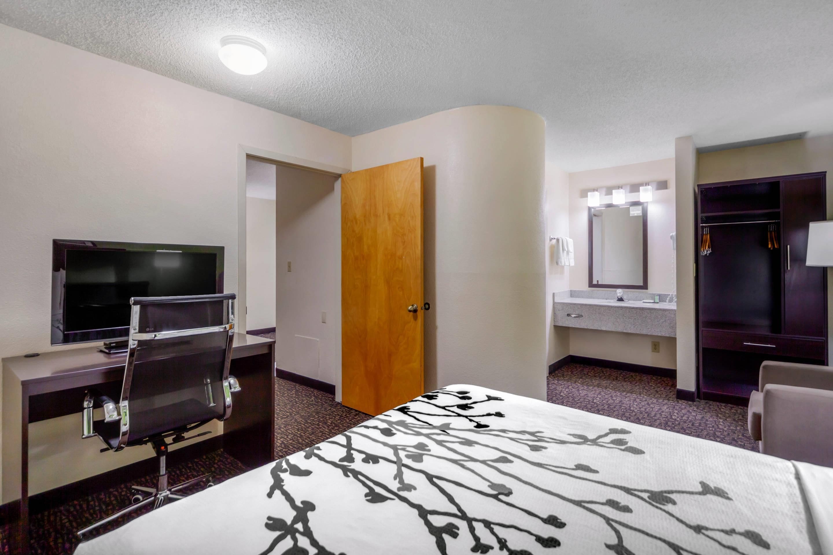 A hotel room with a bed, a desk with a TV, a wardrobe, and a bathroom with a mirror and sink.