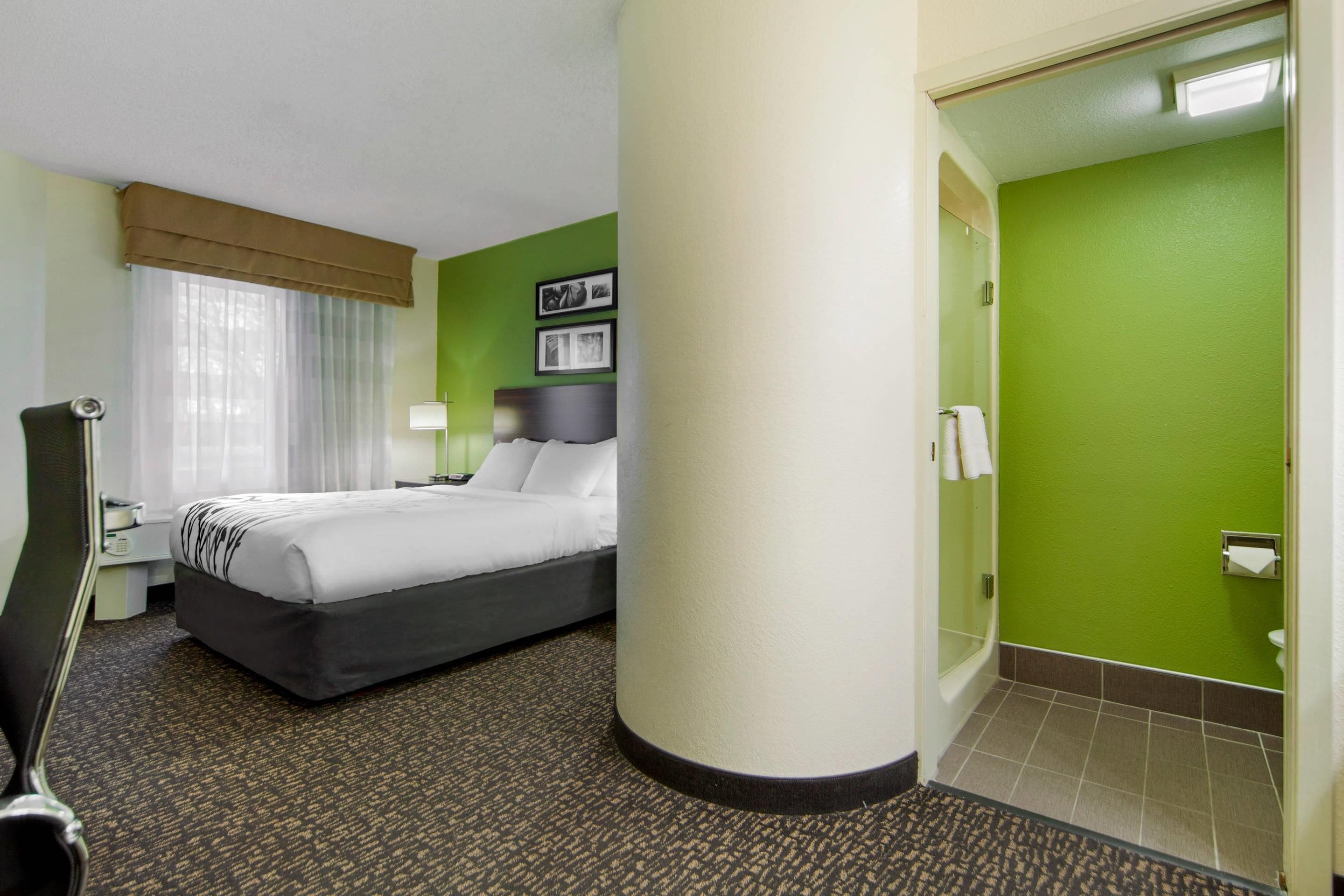 Une chambre d’hôtel comprenant un lit, un bureau, une chaise, une fenêtre avec des rideaux, un mur vert et une salle de bain avec des toilettes et un lavabo.