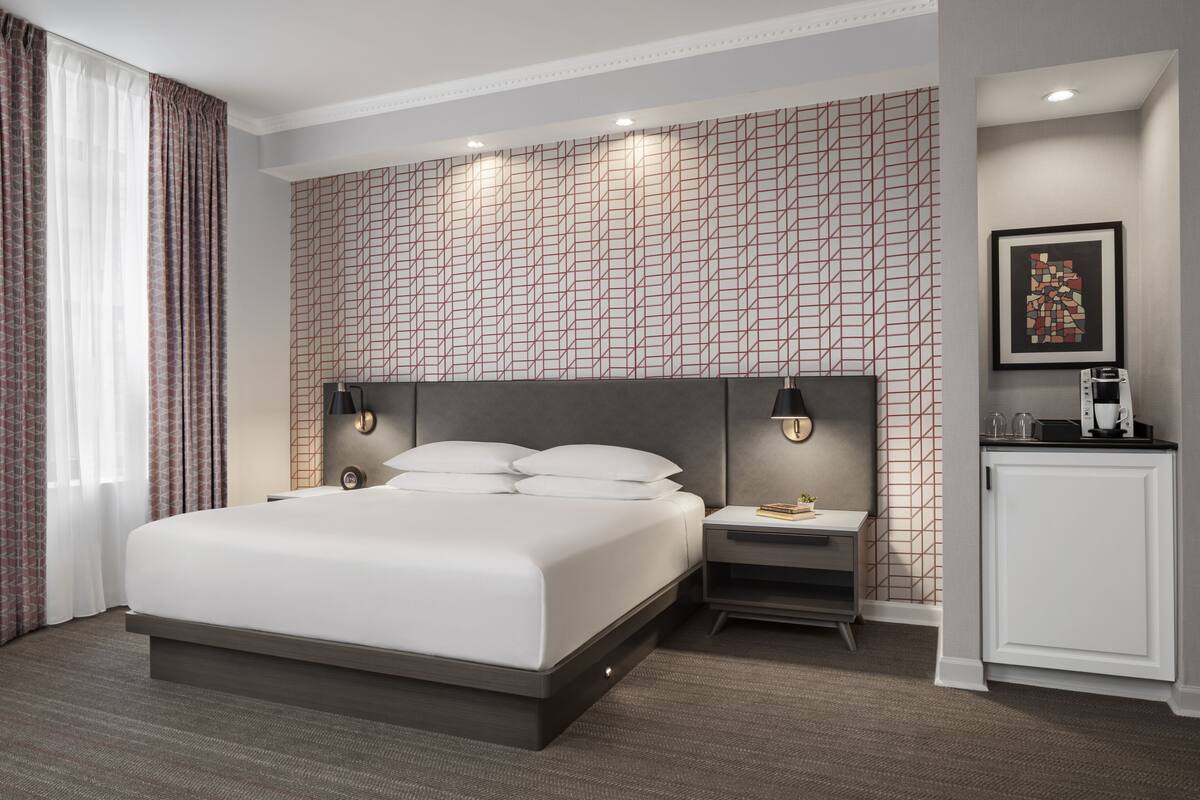 Une chambre d’hôtel moderne avec un grand lit, une table de chevet avec une lampe, une petite armoire et un mur orné d’un motif géométrique.