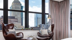 Une chambre d’hôtel moderne, dotée de deux fauteuils en cuir, d’une petite table d’appoint et offrant une vue sur le paysage urbain grâce à de grandes fenêtres.