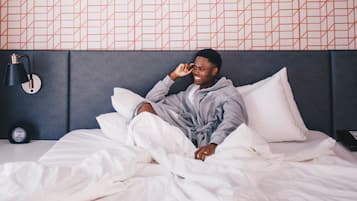 A person lying in bed in a hotel room with a patterned wall and a bedside lamp.