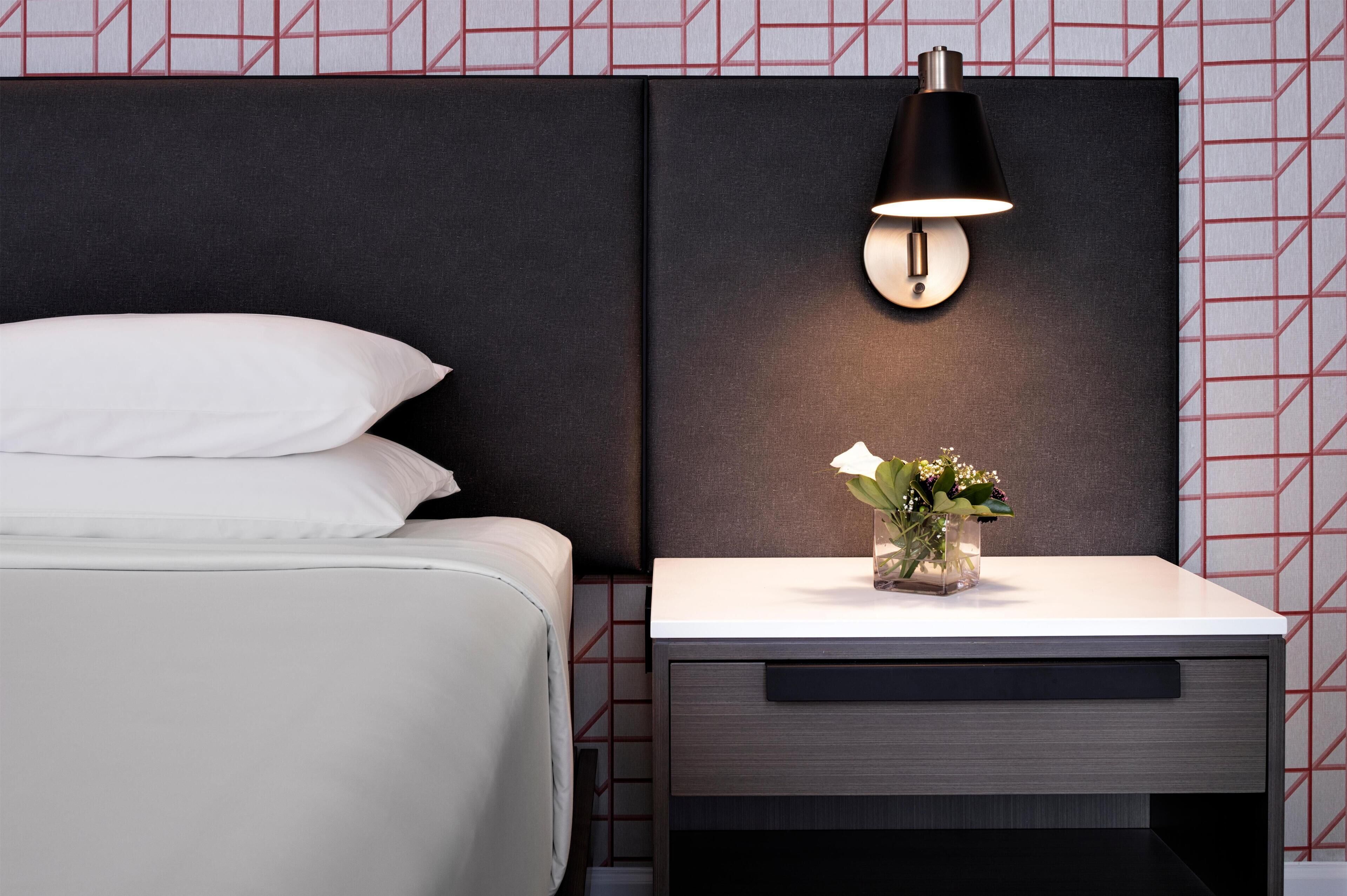 A modern hotel room with a bed, a nightstand with a lamp, and a vase with flowers.