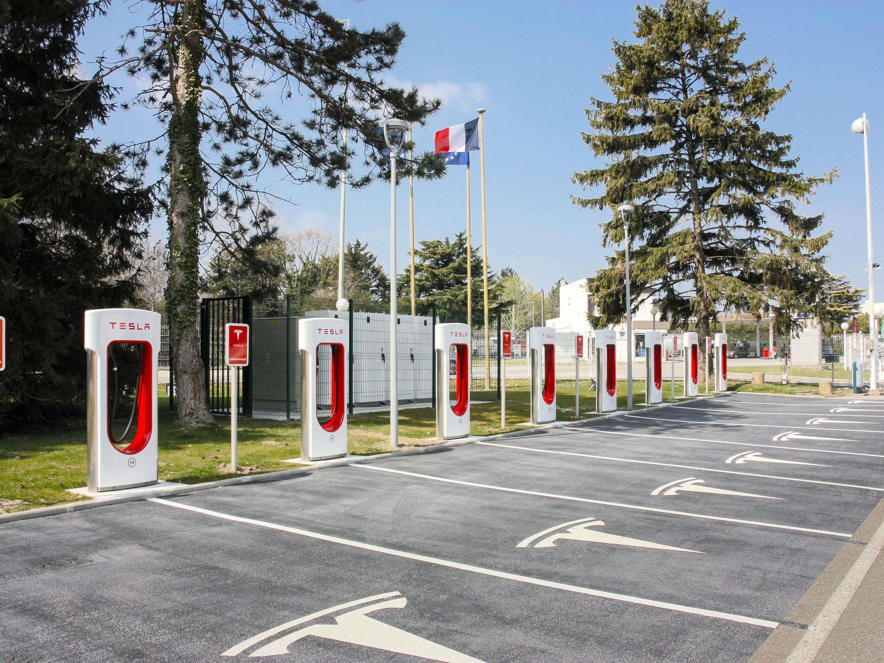 A row of Tesla charging stations with a clear road in front, trees, and a flag.
