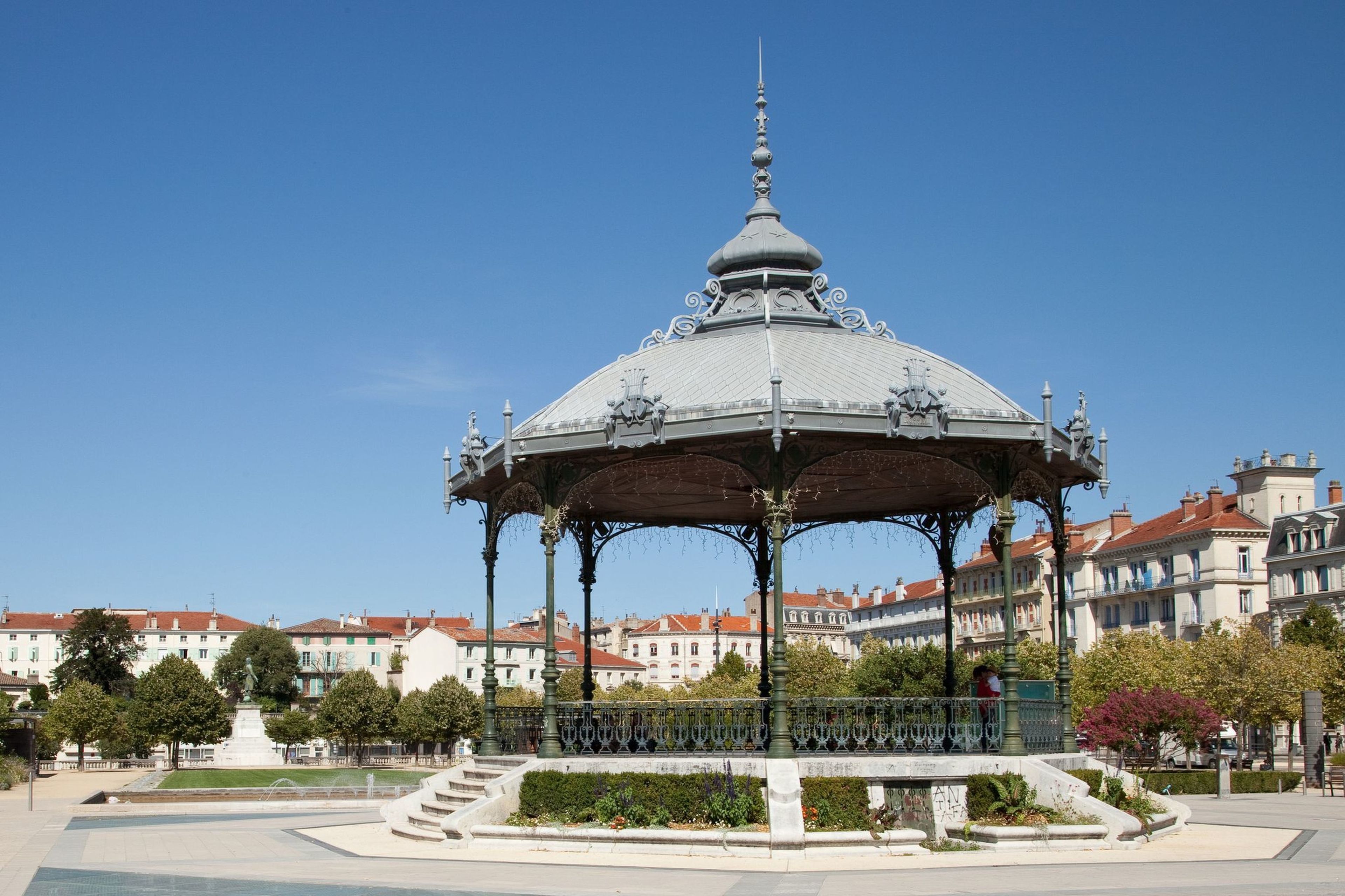 Um coreto com detalhes metálicos elaborados e teto em cúpula, rodeado por um jardim paisagístico e uma fonte, com edifícios históricos ao fundo.