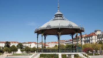 Um coreto com detalhes metálicos elaborados e teto em cúpula, rodeado por um jardim paisagístico e uma fonte, com edifícios históricos ao fundo.