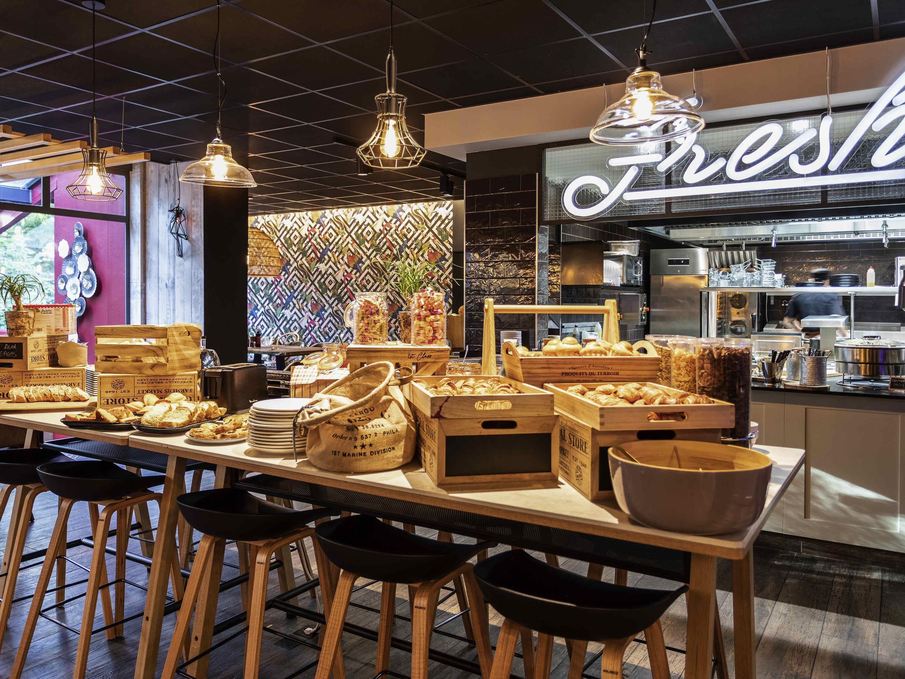 Interior de una cafetería con barra, taburetes de madera, lámparas colgantes y una cocina visible al fondo.