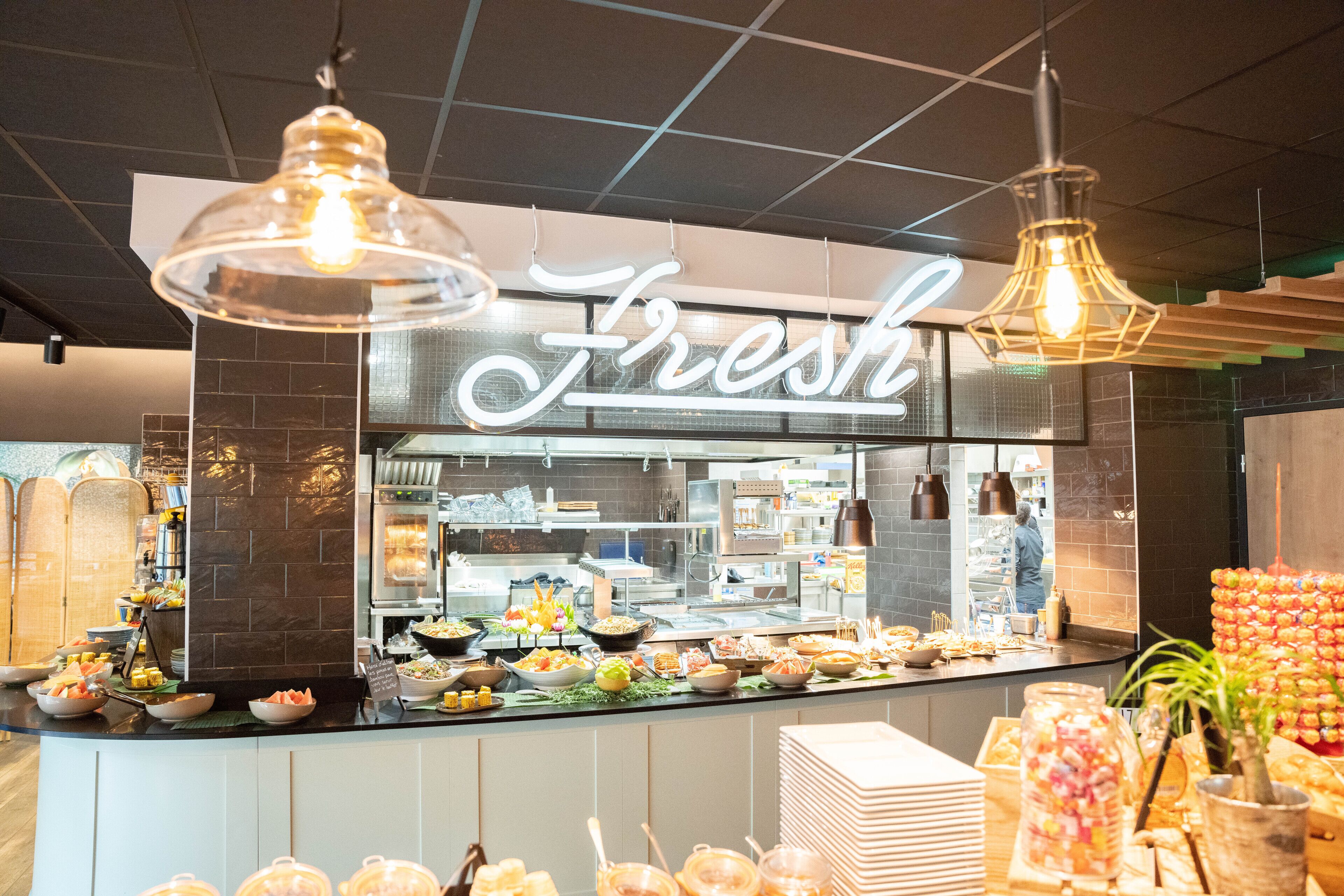 Uma área de buffet com um aviso 'Fresh', diversas opções de comida e luminárias pendentes.