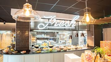 Uma área de buffet com um aviso 'Fresh', diversas opções de comida e luminárias pendentes.