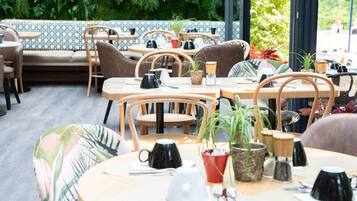Un café avec des tables et des chaises rondes, certaines agrémentées de plantes. On y trouve des tables avec des tasses et des soucoupes, et quelques-unes avec des machines à café.
