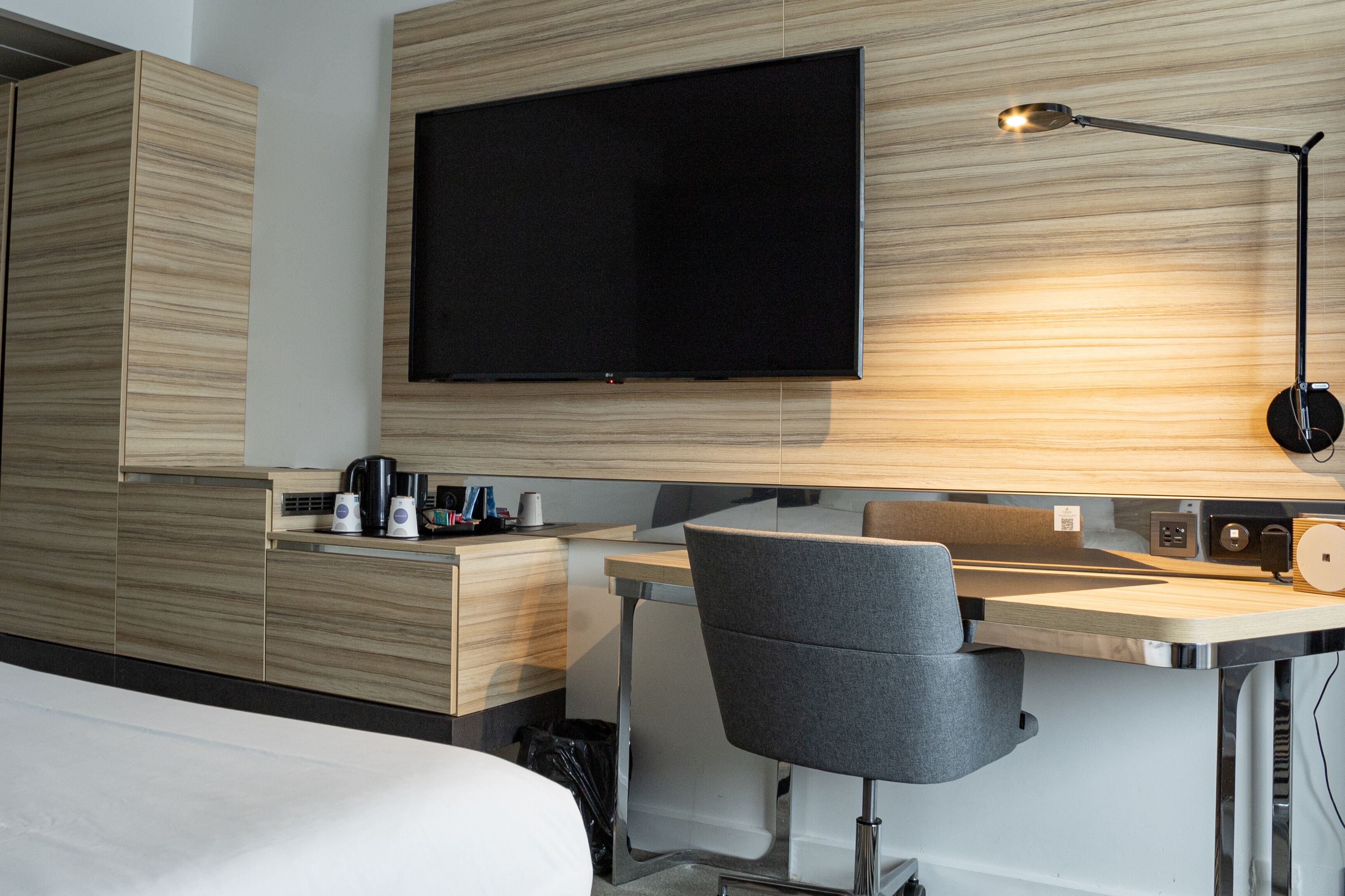 A modern hotel room with a flat-screen TV mounted on the wall, a desk with a chair, and a bedside table with a lamp.