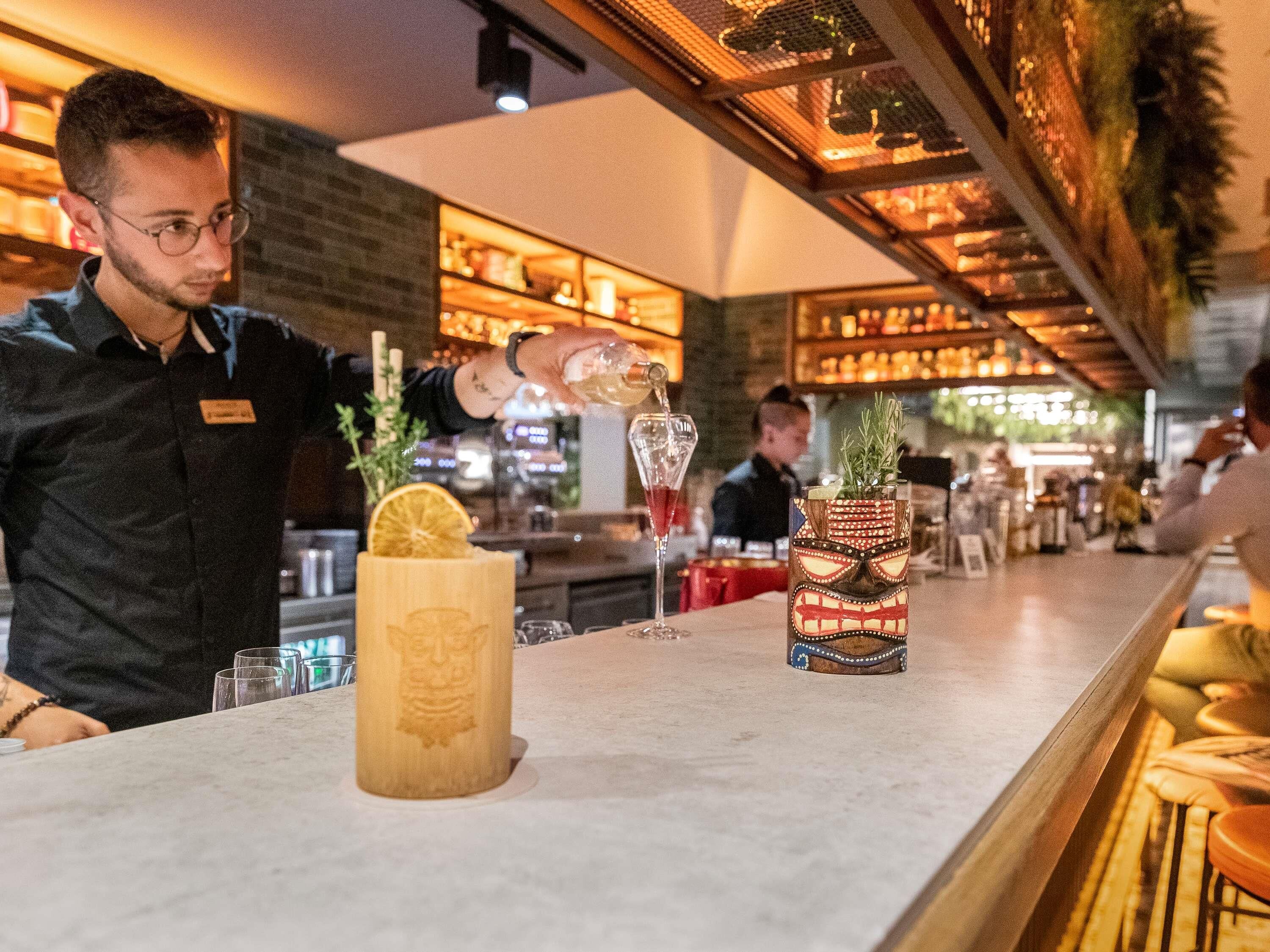 Zona de bar con un cantinero sirviendo una bebida en un vaso. Hay dos jarrones decorativos en la barra, uno con base de madera y el otro con diseño estampado.