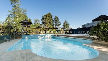 Piscina con sillones de descanso y sombrillas, rodeada de árboles y un cielo despejado.