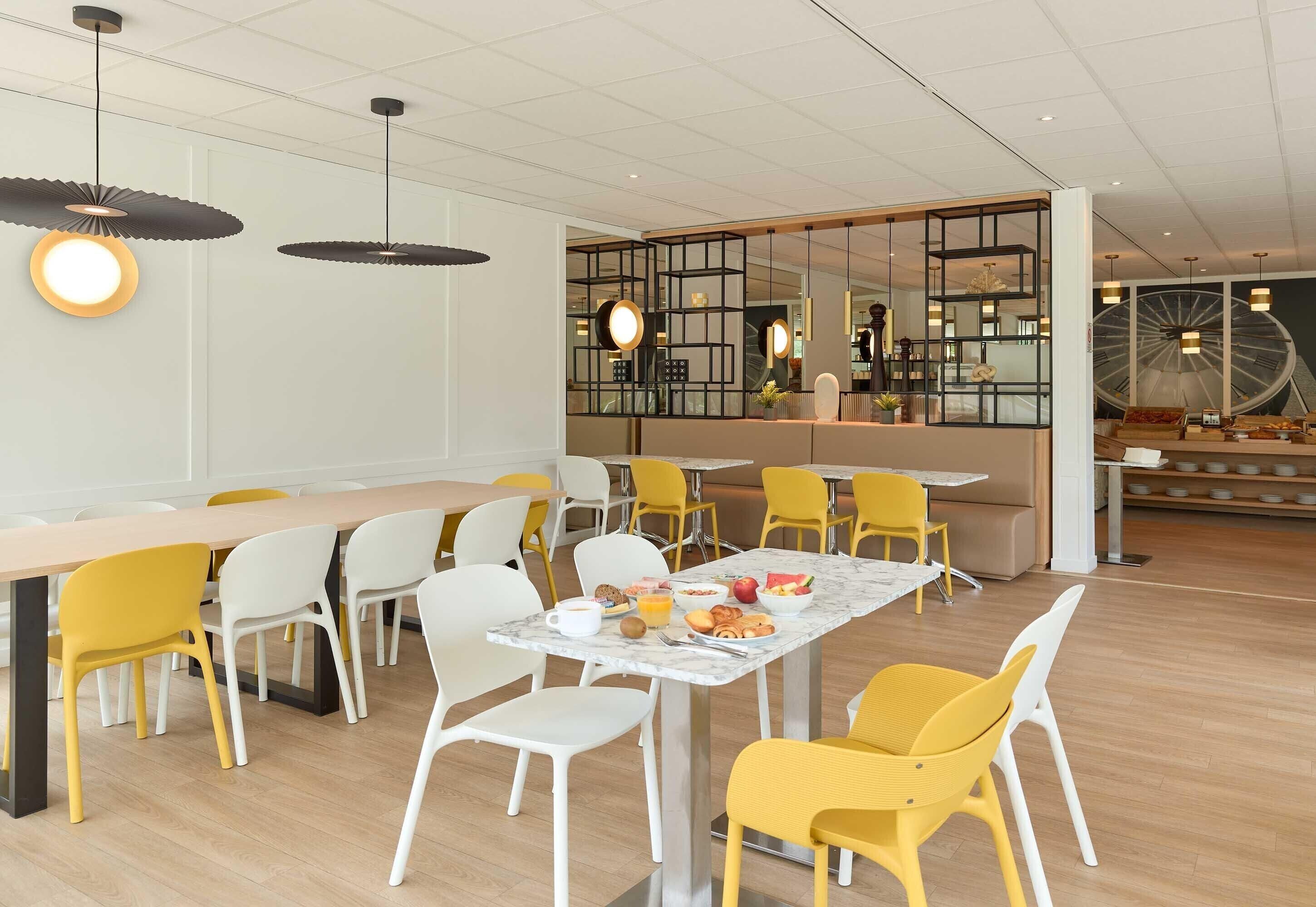 Un espace repas moderne avec des tables et des chaises en bois, un buffet central et une grande fenêtre donnant sur un beffroi.