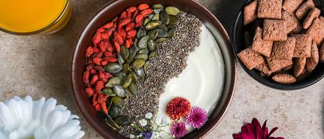 A bowl of yogurt topped with chia seeds, pumpkin seeds, and goji berries, surrounded by flowers and a glass of orange juice.