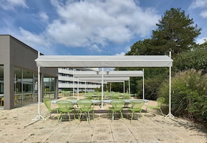 An outdoor seating area with green chairs and tables under a white canopy, adjacent to a building with large windows. - B&B HOTEL Cergy Port (Cergy)