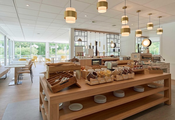 A café with a wooden counter displaying food items, a seating area with tables and chairs, and a large window offering a view of greenery. - B&B HOTEL Cergy Port (Cergy)