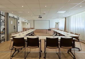 A conference room with a long table, chairs, a projector screen, and a large window with curtains.