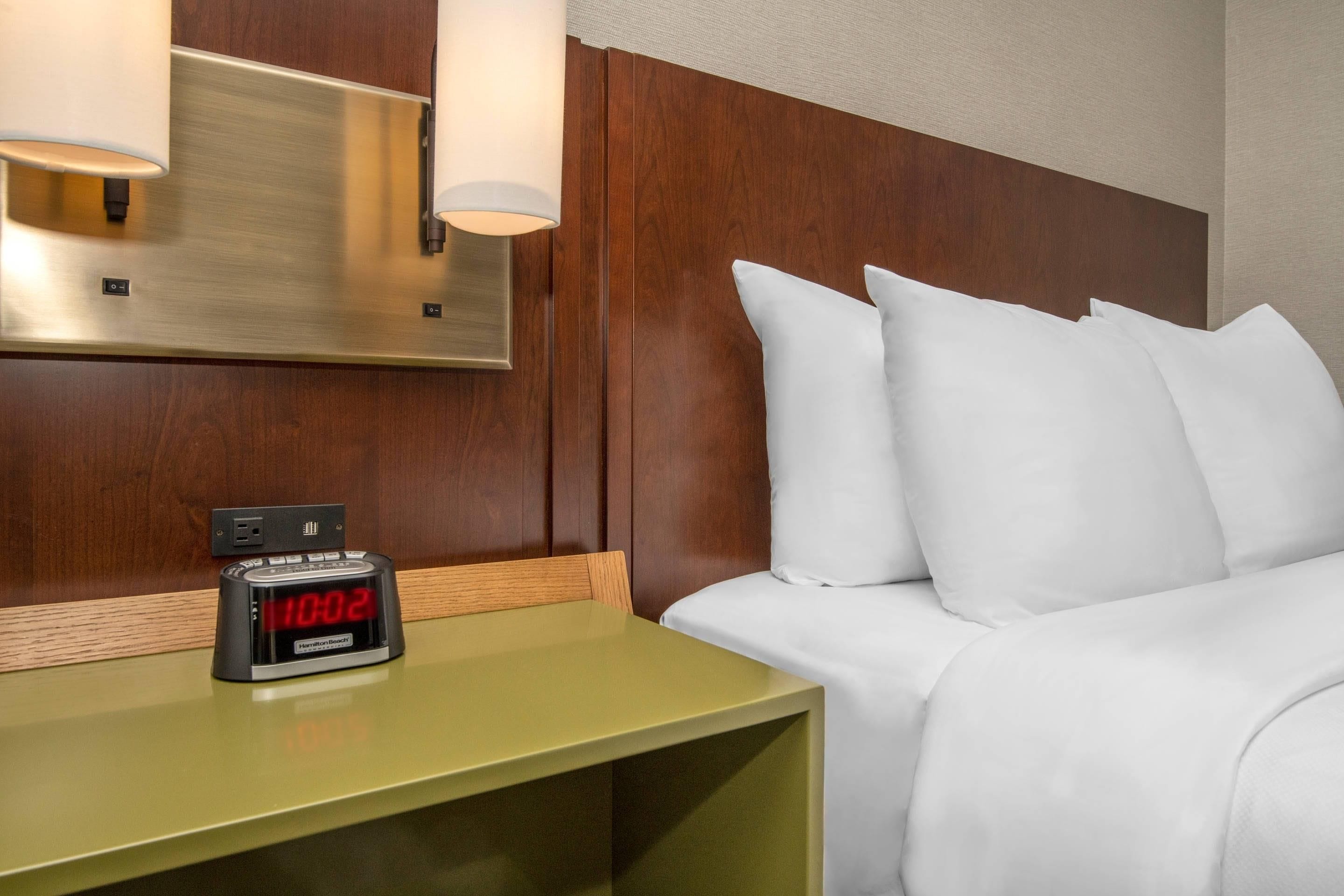Une chambre d’hôtel comprenant un lit, des oreillers, une table de chevet avec une horloge numérique et un luminaire fixé au mur.