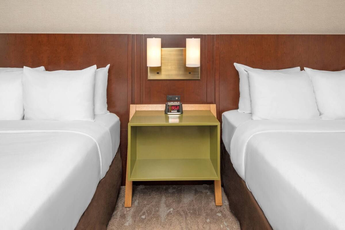 A hotel room with two beds, a small green nightstand with a clock, and wall-mounted lamps.