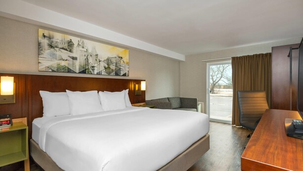 Ein Hotelzimmer mit einem großen Bett, einem hölzernen Kopfteil, einem Gemälde über dem Bett, einem Schreibtisch mit Stuhl und einem Fenster mit Vorhängen.