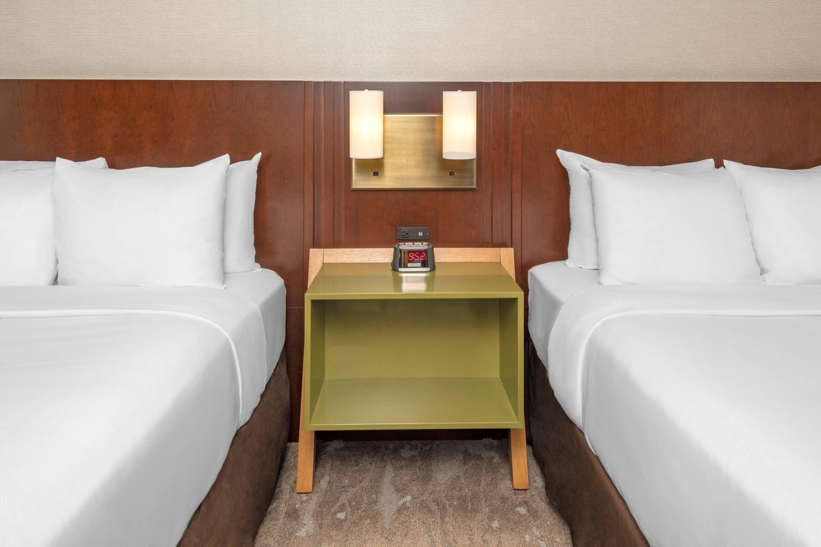 A hotel room with two beds, a nightstand with a clock, and a wall-mounted light fixture.