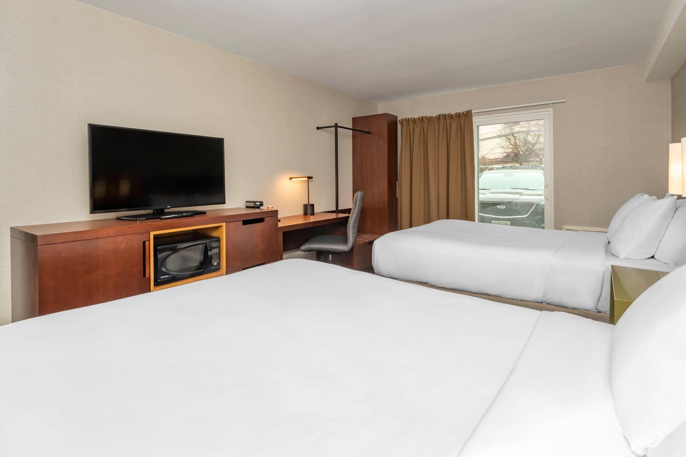A hotel room with a flat-screen TV, a desk with a lamp, a microwave, two beds, and a window with curtains.