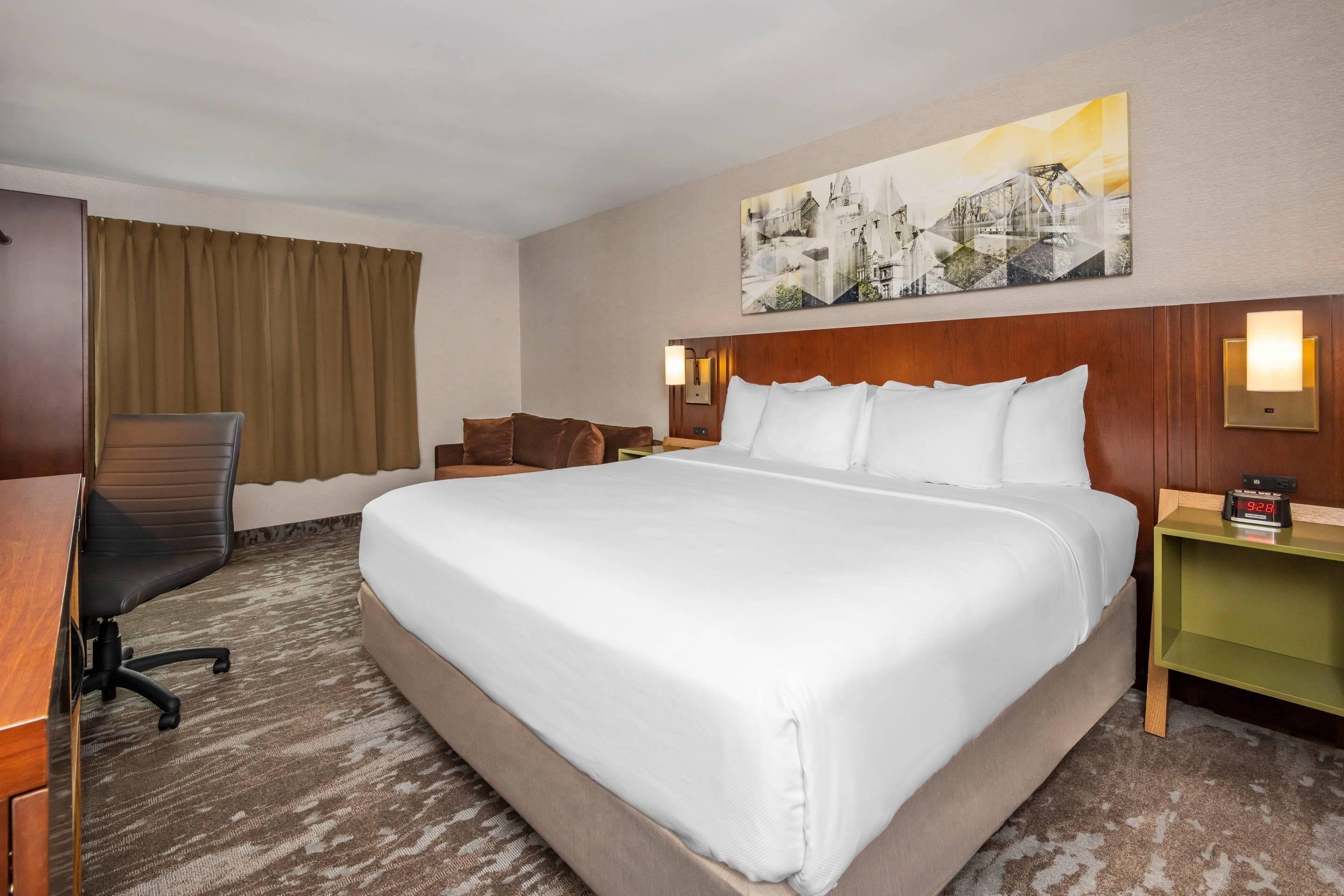 A hotel room with a large bed, a desk with a chair, a nightstand, and a wall-mounted clock.
