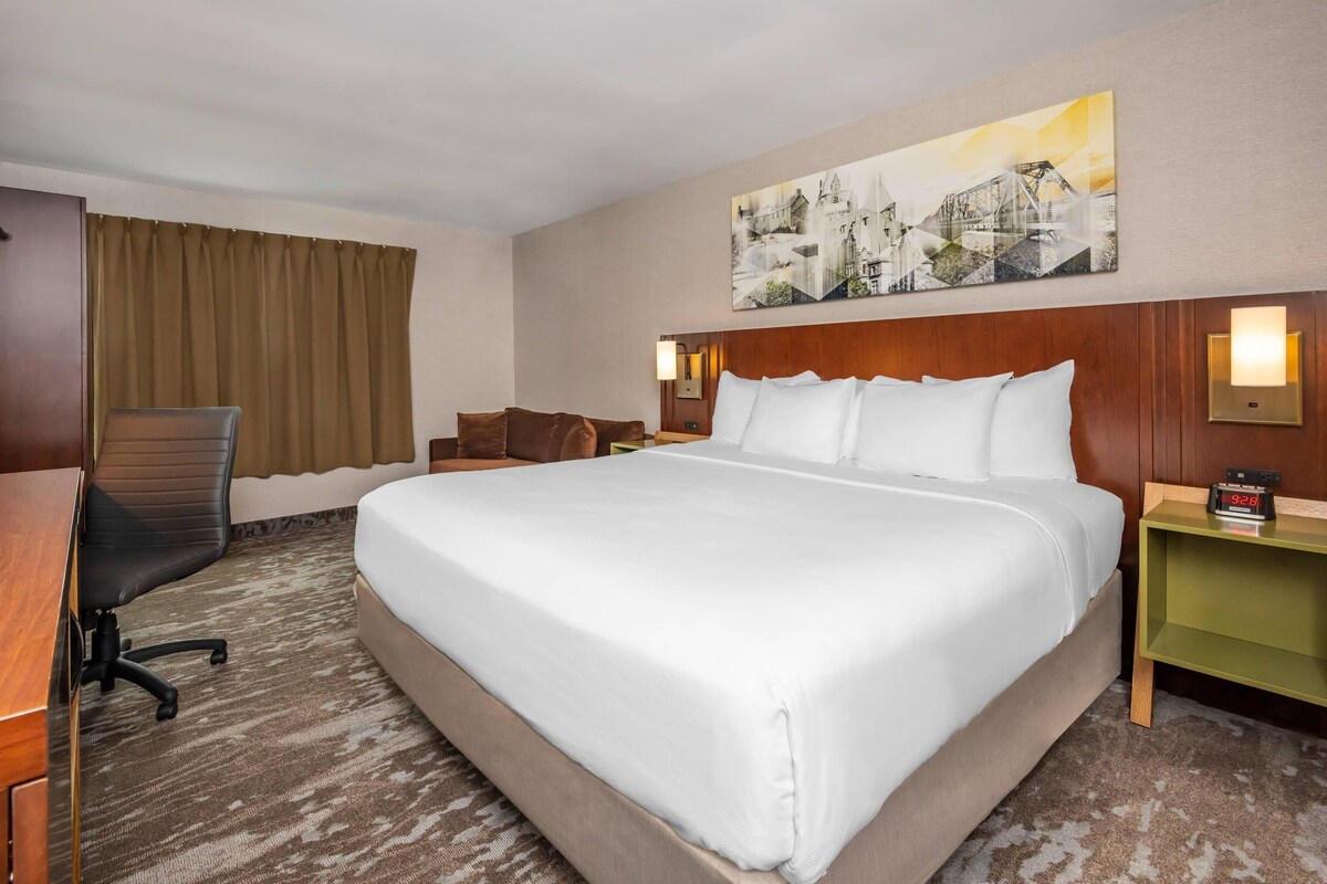 A hotel room with a large bed, a desk with a chair, a nightstand, and a wall-mounted clock.