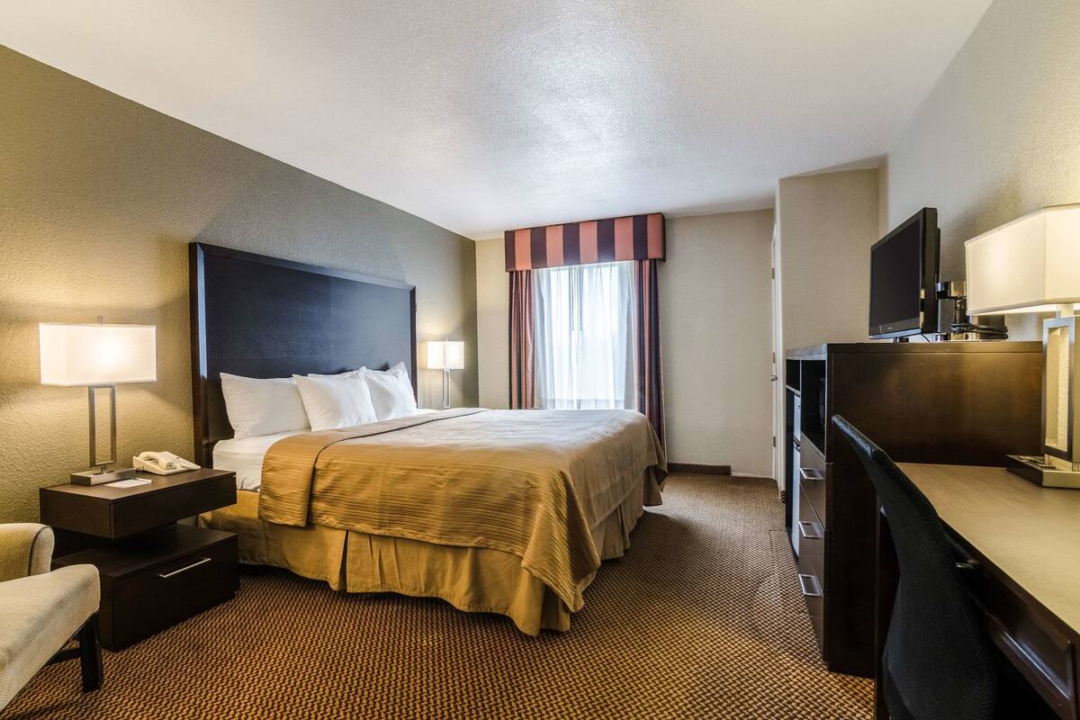 A hotel room with a large bed, bedside tables, a desk with a computer, a TV, and a window with curtains.