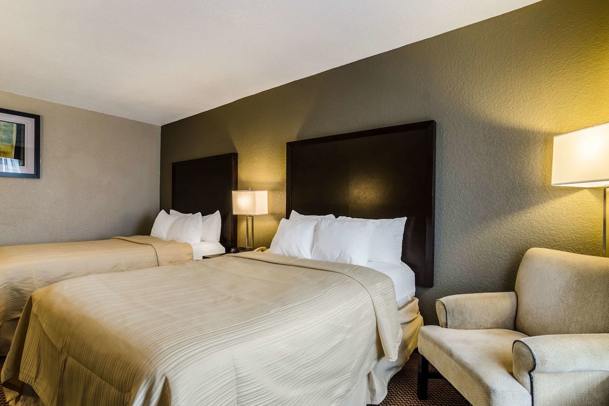 A hotel room with two beds, a chair, and a nightstand with a lamp.
