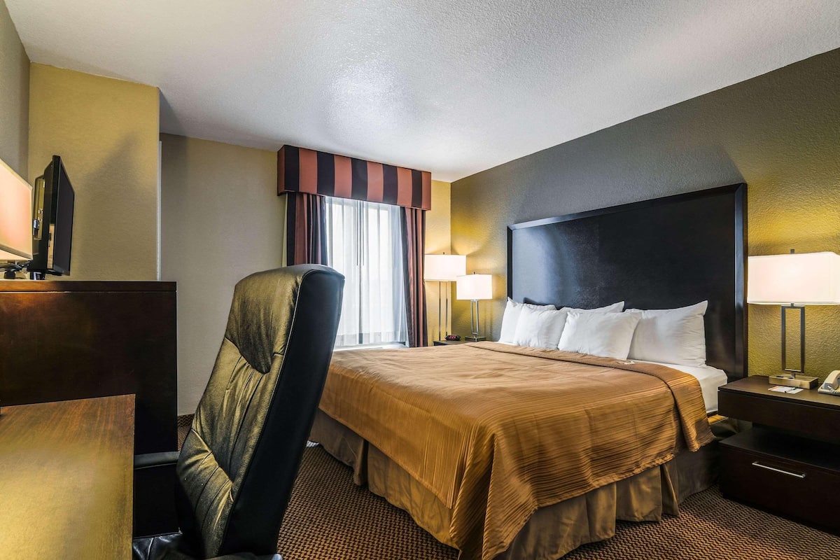A hotel room with a large bed, a desk, a chair, and a nightstand with a lamp.