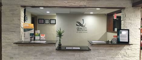 Un comptoir d’accueil avec un mur en pierre en arrière-plan, le logo de « Quality Inn & Suites by Choice Hotels » et une petite plante en pot.