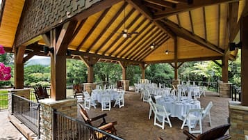 An outdoor covered patio with tables and chairs, stone flooring, and wooden beams.