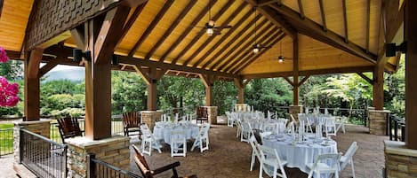 An outdoor covered patio with tables and chairs, stone flooring, and wooden beams.