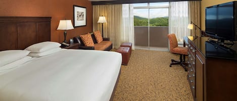Habitación de hotel con una cama grande, un escritorio con lámpara, una silla, un televisor y un balcón con vistas a las montañas.