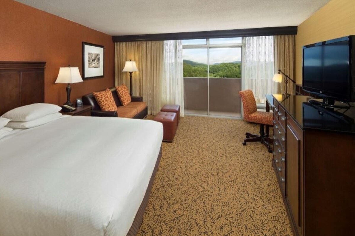 A hotel room with a large bed, a sofa, a chair, a TV, and a balcony with a view.