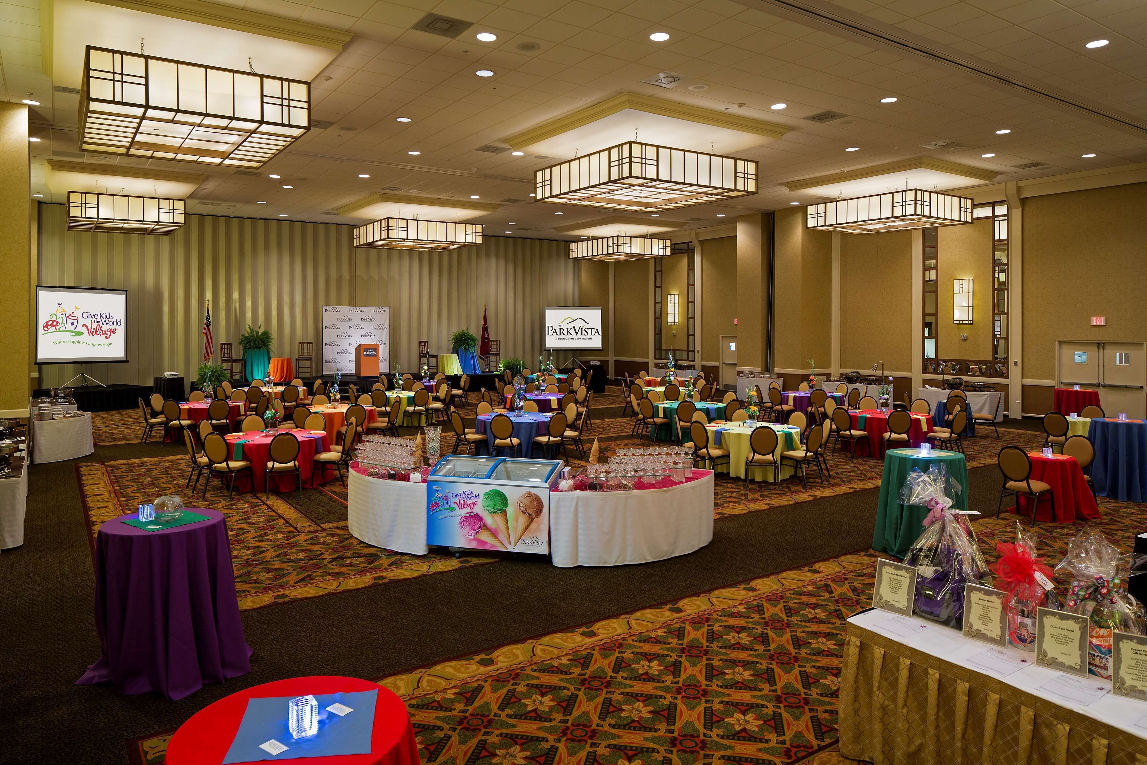 A large conference room with multiple tables and chairs, a stage with a presentation screen, and a carpeted floor.